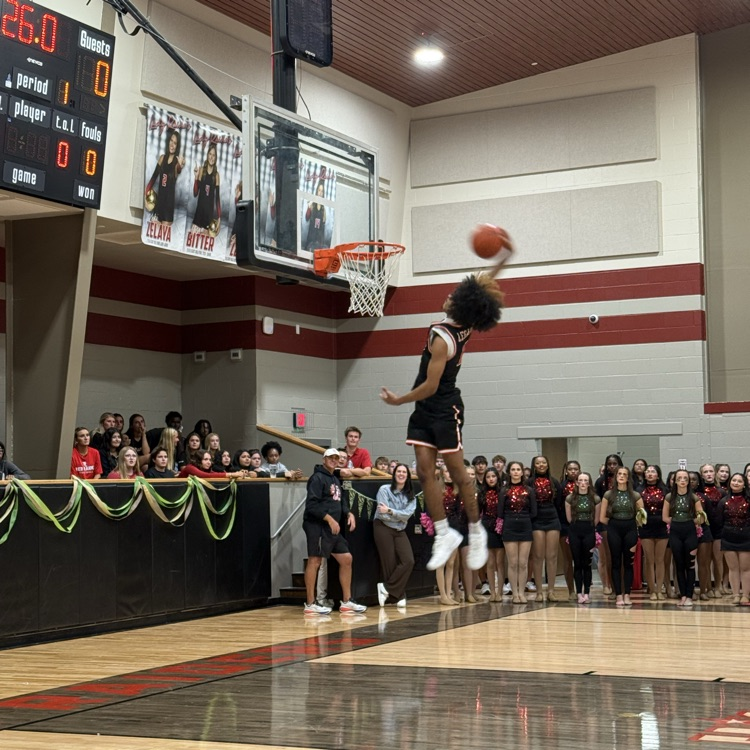 Indoor basketball court with a player jumping to dunk the ball. Audience on benches watching. Scoreboard on wall.