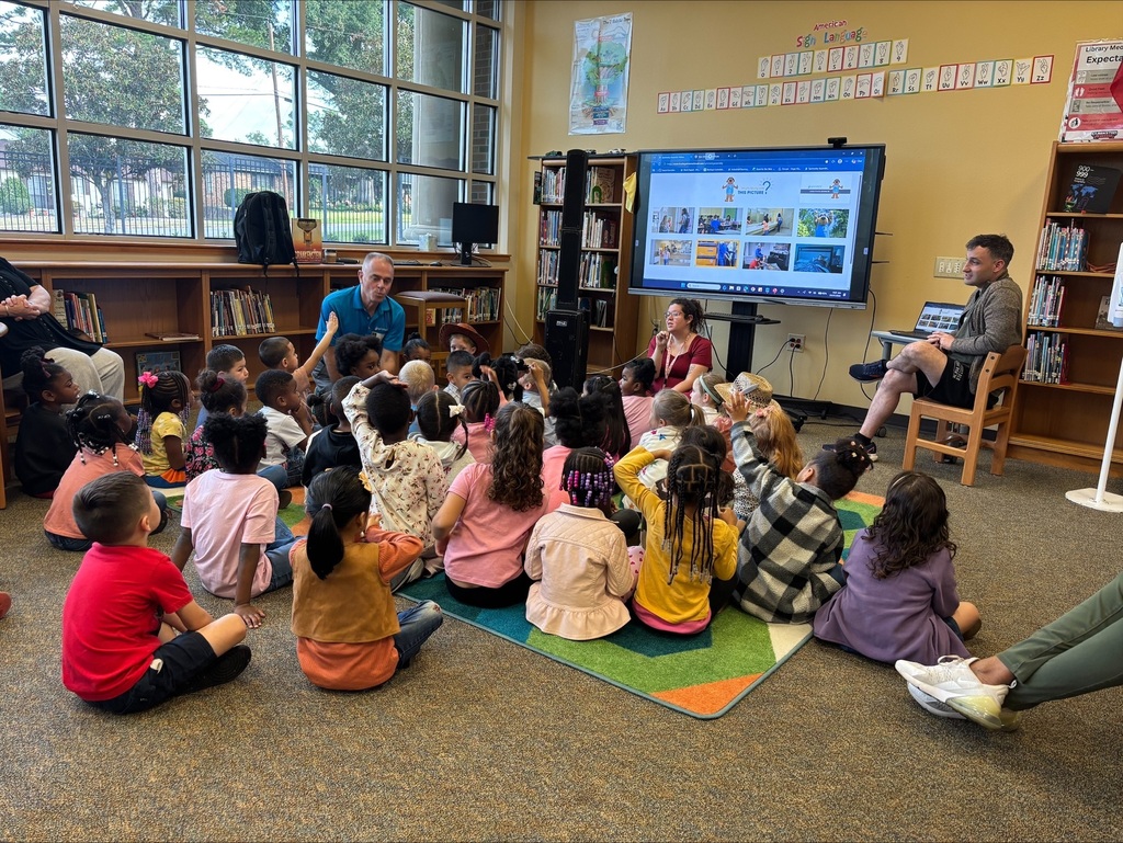A group of children sit around on a rug in a classroom. A man stands in front of a screen.