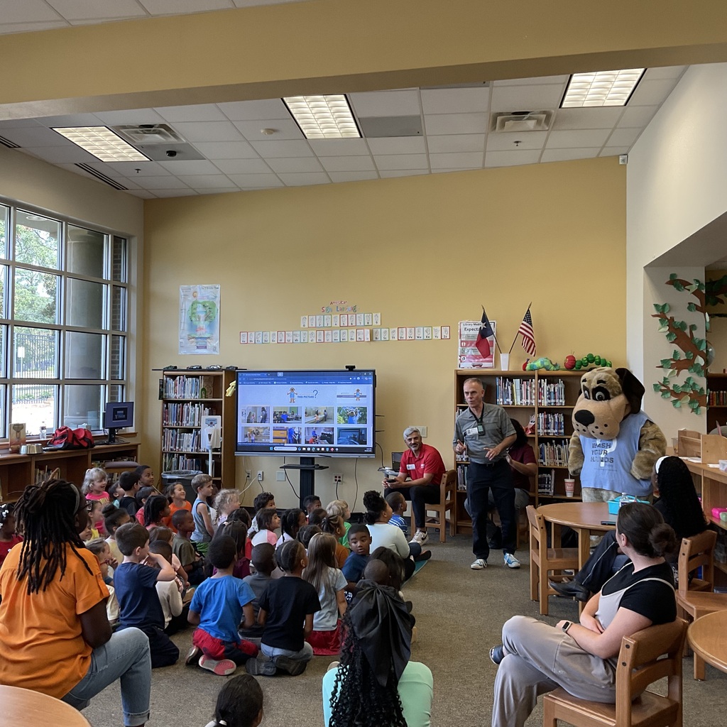 Students gathered in a room with bookshelves, listening to a man with a dog mascot. A large screen shows images.