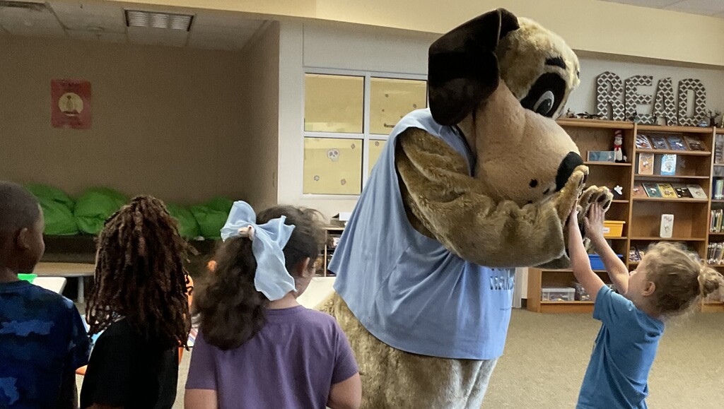 Children in a classroom interact with a mascot resembling a dog in a blue t-shirt.