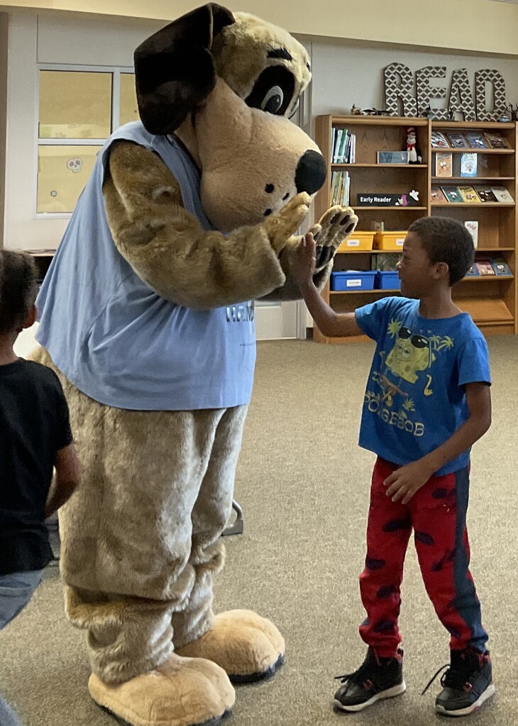 Mascot dog in light blue shirt shaking hands with a boy in red pants. Library background.