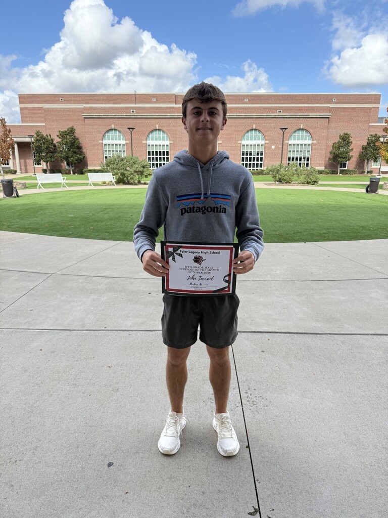 Teen in gray hoodie with Patagonia logo holds framed certificate in front of building with arched windows.