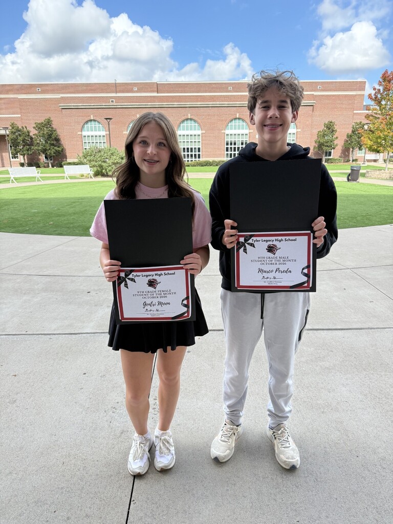Two children stand next to each other holding certificates. The girl wears a pink shirt and black skirt. The boy wears a black hoodie and white pants.