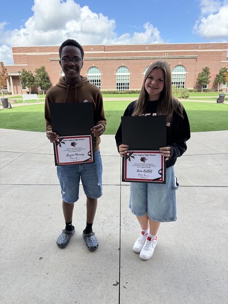 Two students stand in front of a building with certificates in black frames. One holds a certificate in red ribbon.