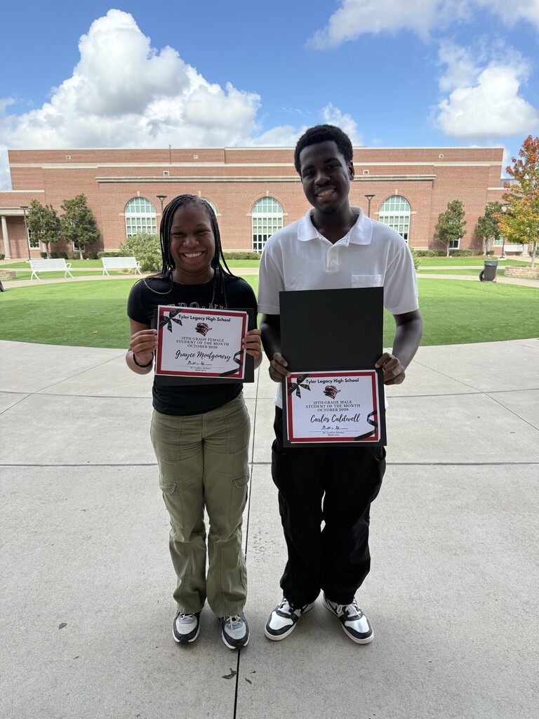 Two students hold framed certificates outside a brick school building on a sunny day.