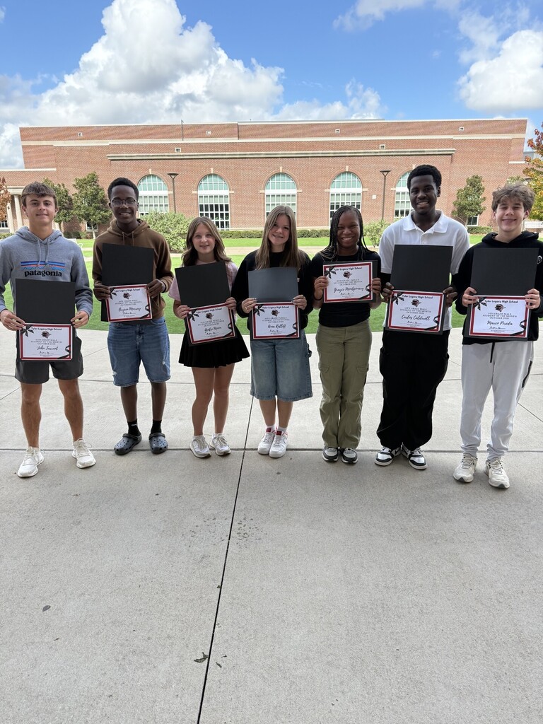 Students pose for photo, holding certificates, in front of a brick building with trees and a cloudy sky.