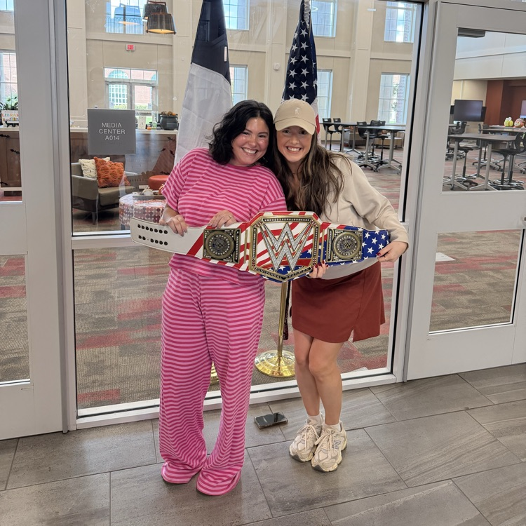 Two women in front of glass doors at a school, one holds a belt with USA flag.