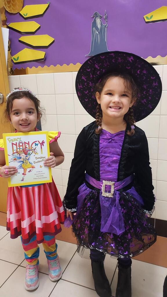Two girls in Halloween costumes, one holding a book, standing in a classroom.