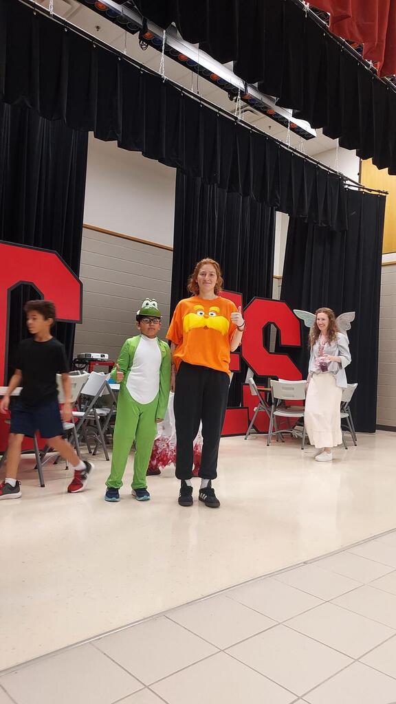 People in costumes on a stage; a person in orange stands with two children, one in green.