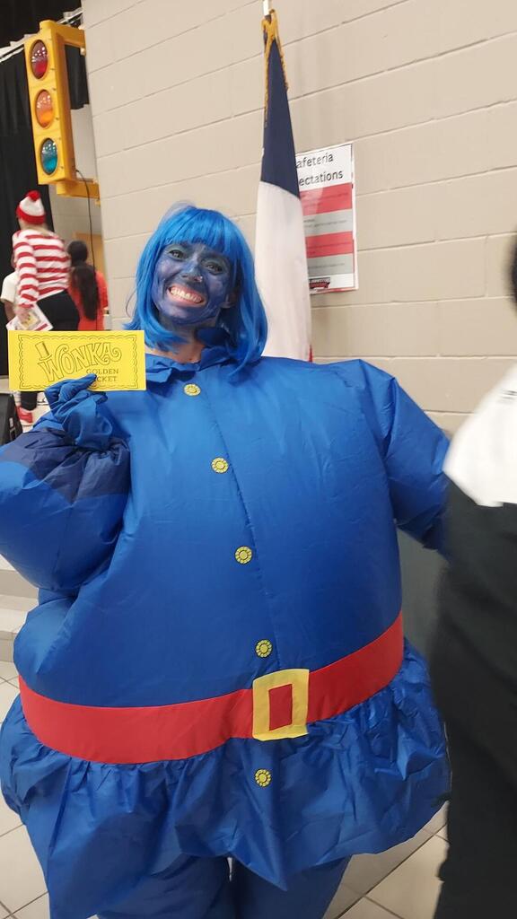 Person dressed in blue with face painted blue, holding yellow paper, standing in front of a flag.