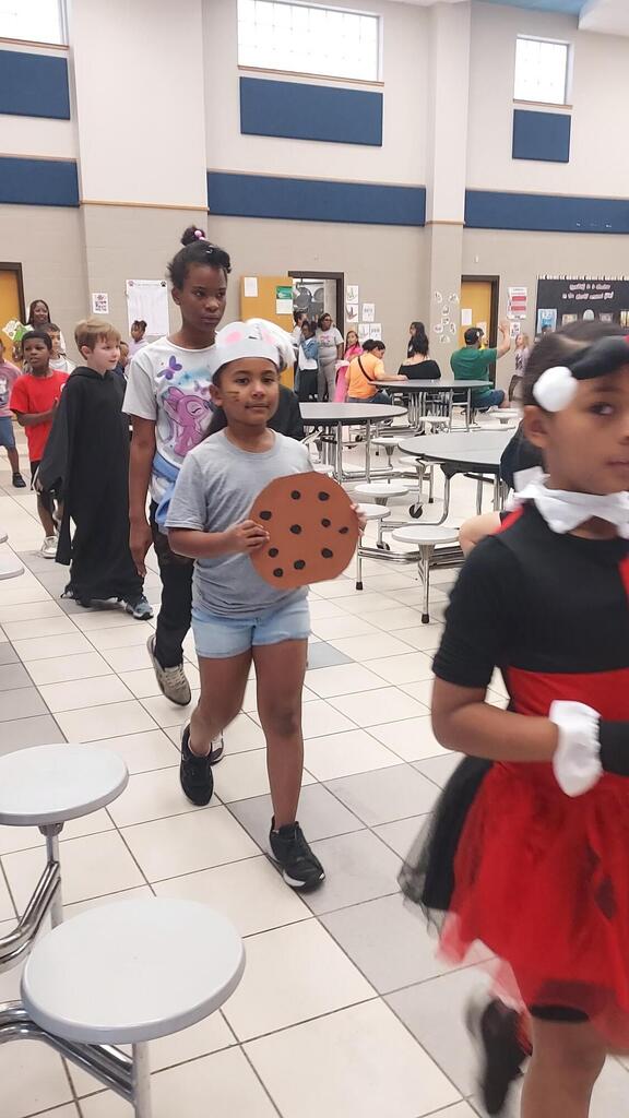 Students in costumes walking through a cafeteria; one holds a large cookie. Tables and chairs in the background.