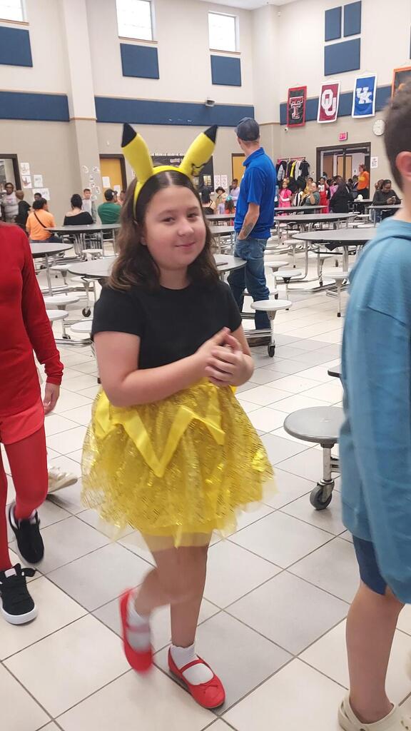 A young girl wearing a yellow skirt and red shoes poses in a cafeteria with tiled floors.