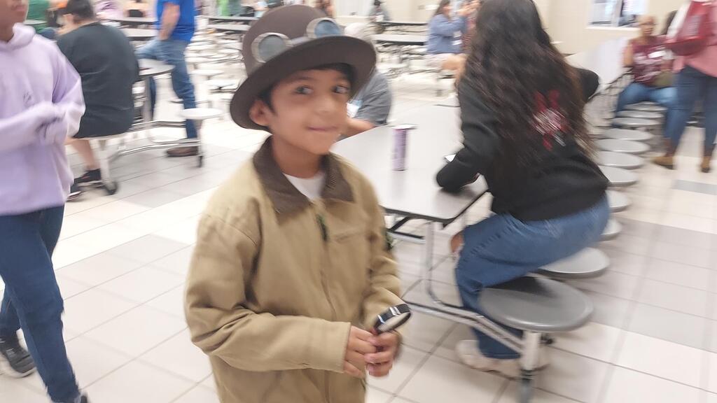 A young boy wearing a brown hat holds a metal object in a school cafeteria.