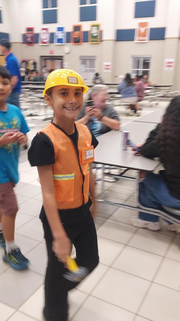 Young boy in yellow hard hat and safety vest stands in hallway, smiling. Other children and adults sit at tables in background.