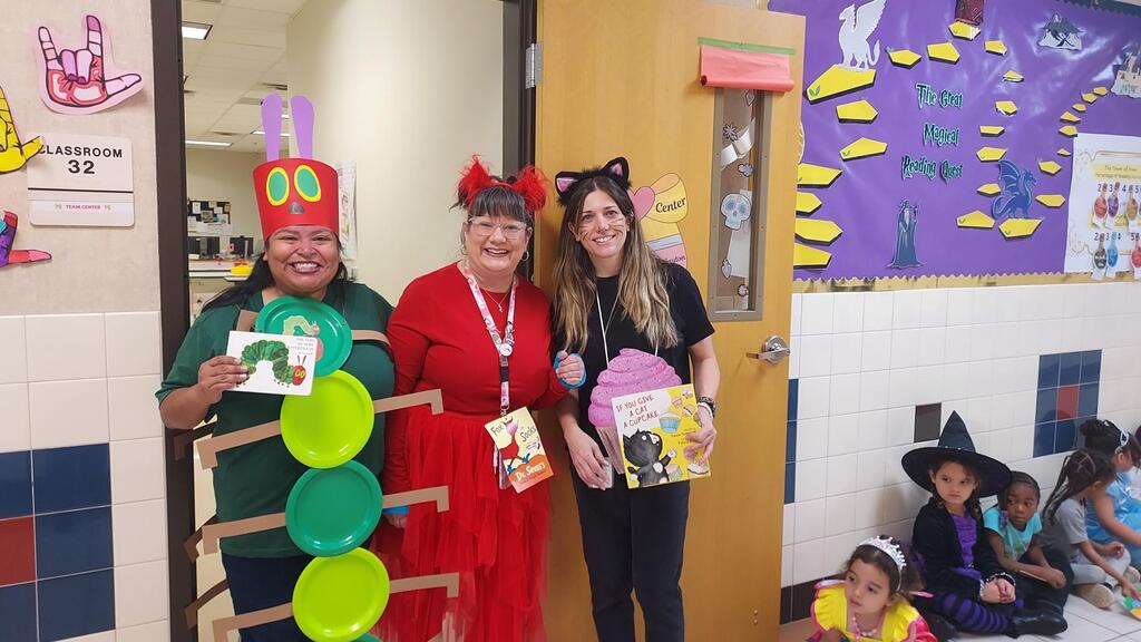 Three adults in costumes, holding books, stand near a door. Children in costumes sit on the floor.
