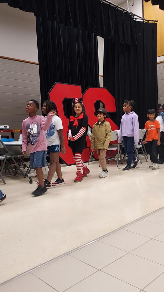 Several children in costumes line up on a stage with a large red "SC" backdrop.