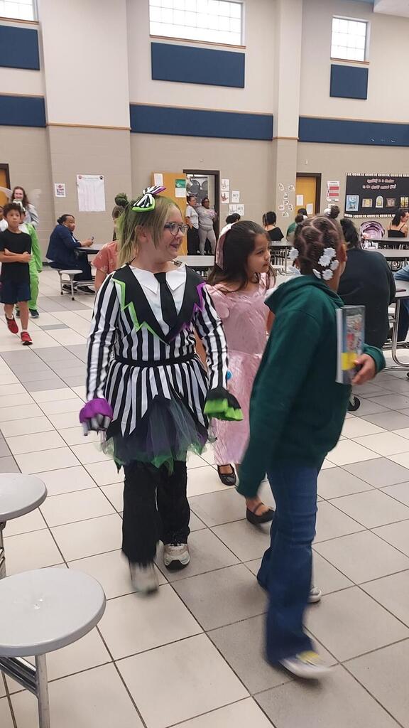 Students in costumes walk in a school hallway with tiled floor and round tables. Some people sit at tables.