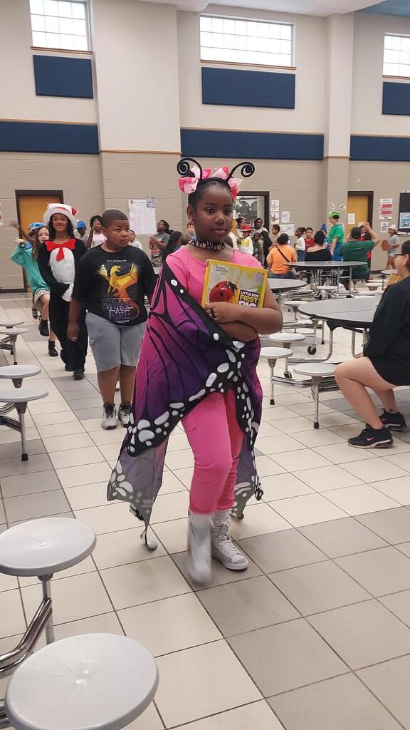 A girl in costume stands in a school hallway with other students and adults.