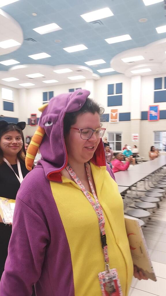 Adult in a purple and yellow costume holds a book, standing near a person in black.