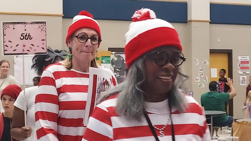 Two people in matching red and white striped outfits and hats smile at the camera, standing in a school hallway.