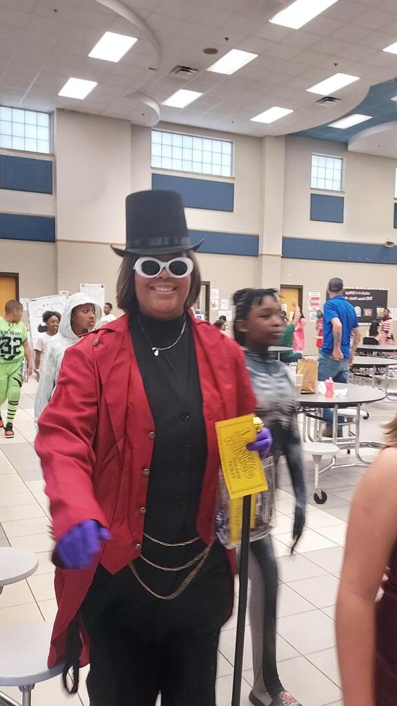 A person in a red coat and top hat holding a yellow paper stands in a room.