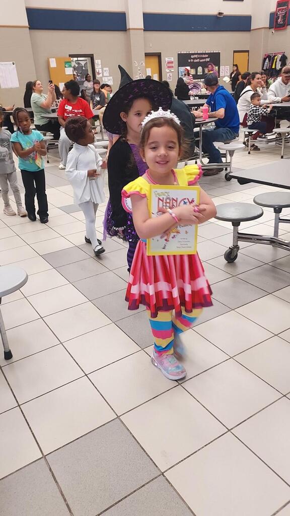 Child in colorful dress holding book, standing in school hallway with tiled floor and people.