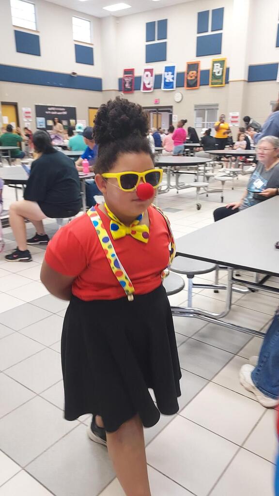 A girl in a red shirt, black skirt, and yellow suspenders with a red clown nose poses in a dining hall.