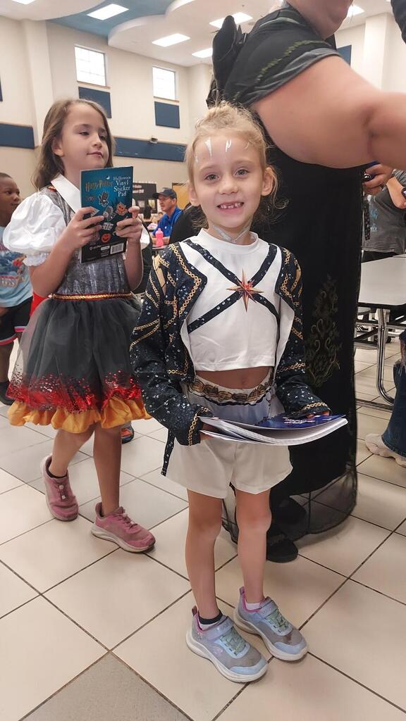 Two children stand in a tiled room; one holds a shoe, and the other holds a tablet. Behind them, a person stands, and others are visible in the background.
