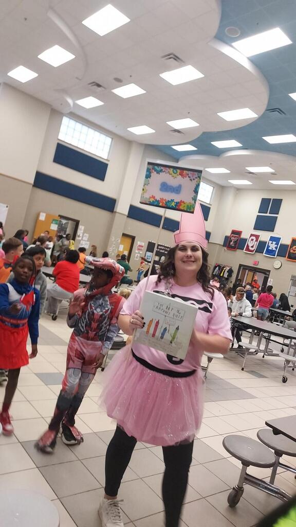 Person in pink holds book, surrounded by kids in costumes in a room with tables and chairs.