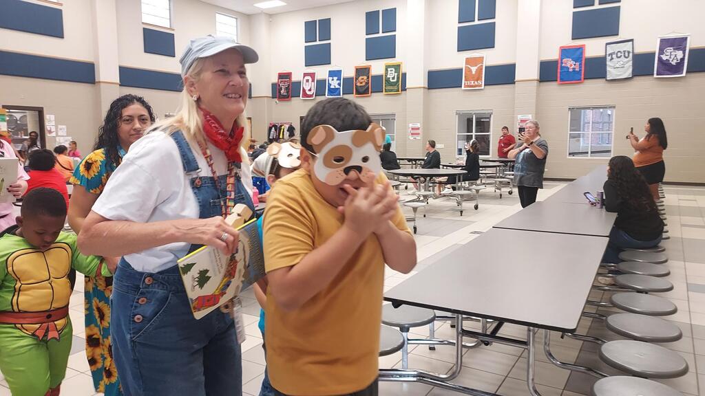 A woman and boy in costumes in a school cafeteria with students, tables, and flags on the walls.