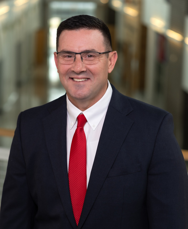 Caucasian man with short dark hair and glasses wearing a red tie and dark suit jacket