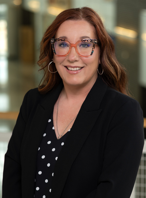 Caucasian woman with brown hair and glasses wearing polka dot shirt and dark jacket
