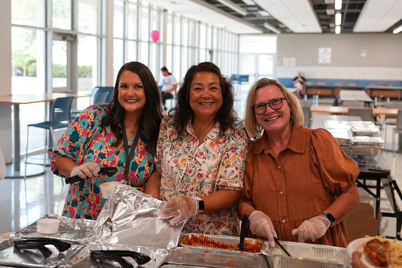 three women standing behind a table with food to serve