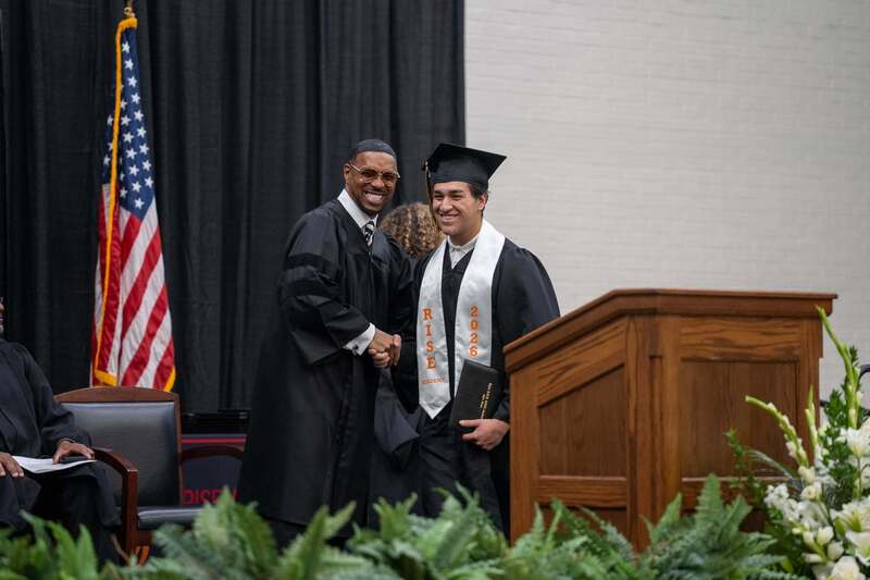 African American man wearing graduation gown shakes hand of teen boy wearing graduation gown on stage
