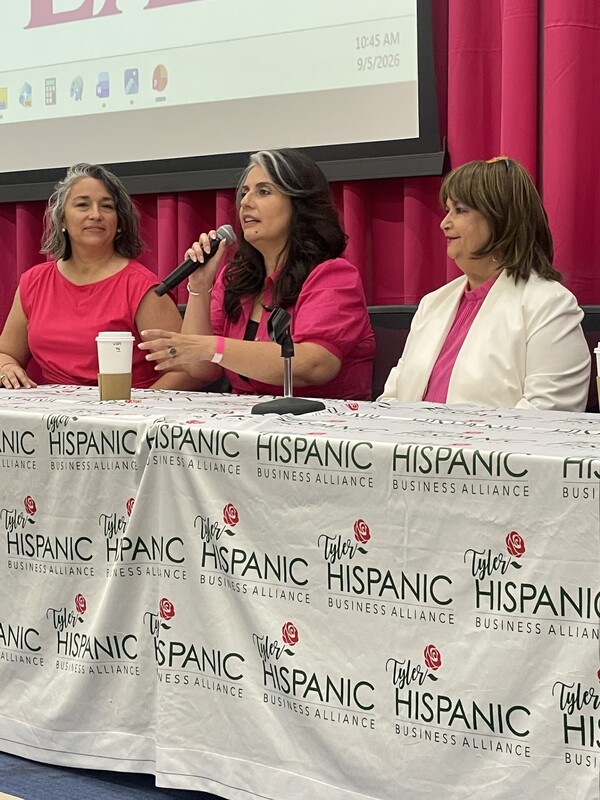 three Hispanic women sitting at table, one holding microphone as she talks