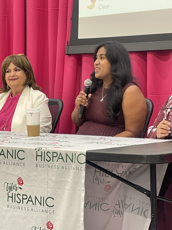 two Hispanic women sitting at table, one holding microphone as she talks