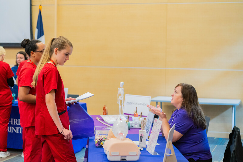 woman sitting at table talking to teen girl as she looks at CPR mannequin
