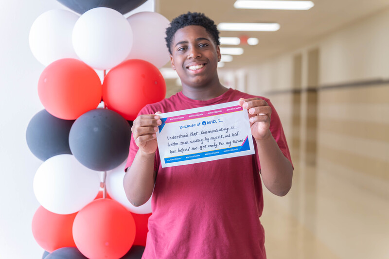 teen boy holding certificate in school hallway, standing next to balloons