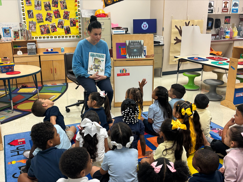 Assistant Principal Mrs. Bernardino holds up a book about teachers as Bonner students sit on the classroom rug, with one student raising a hand.