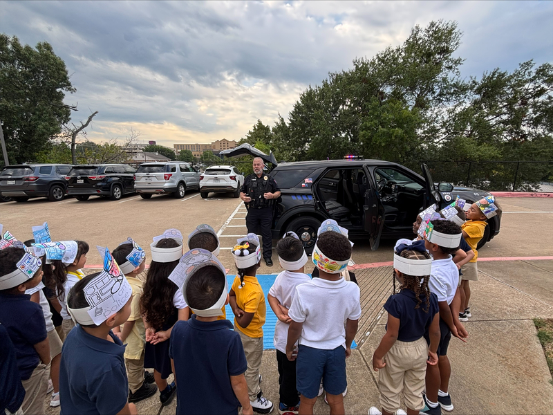 Officer Johnson speaks with Bonner students gathered outside beside a police vehicle. Students wear paper hats decorated with community helper pictures.