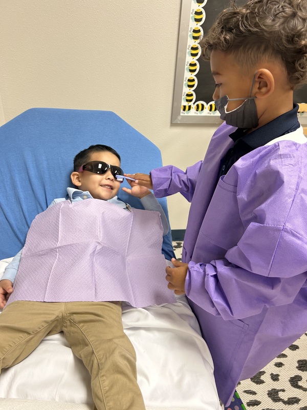 A student wearing a mask and purple gown holds a toothbrush near the mouth of a classmate reclining in a dental chair with protective glasses and a purple bib.