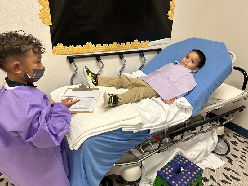 A student wearing a mask and purple gown holds a clipboard beside a classmate reclining in a dental chair with a purple bib.