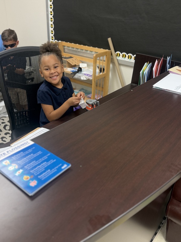 A smiling student sits behind a desk, holding dental supplies beside a model of teeth.