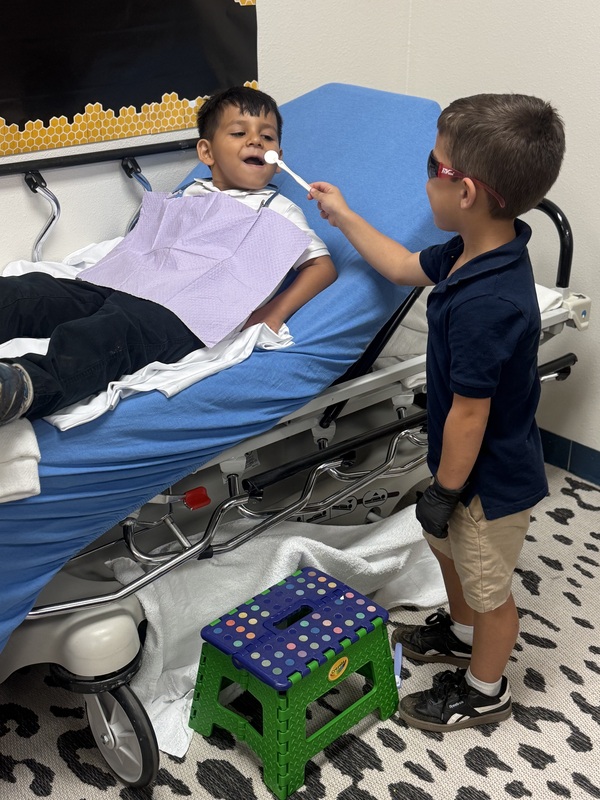 Two students practice a pretend dental exam. One reclines in a dental chair wearing a purple bib while the other holds a dental mirror near his mouth.