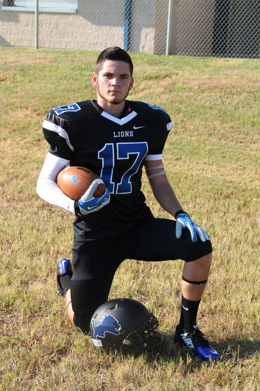 teen boy in his high school football uniform kneeling in grass holding football