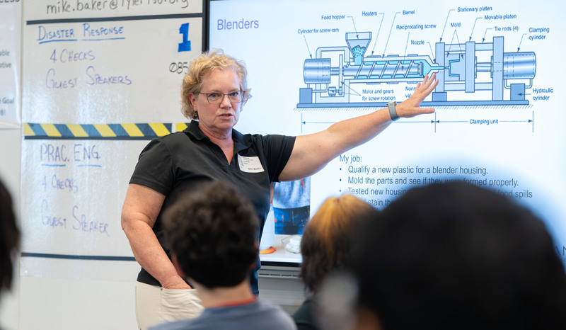 woman pointing at diagram on board in front of high school classroom