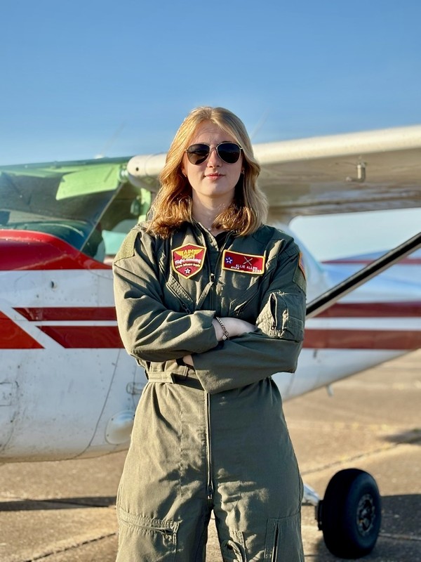 teen girl with blond hair wearing jumpsuit standing in front of airplane