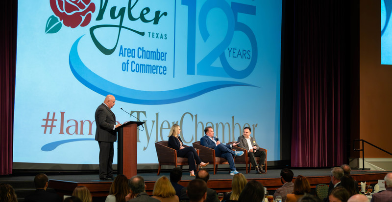 two men and a woman sit on stage with another man behind microphone