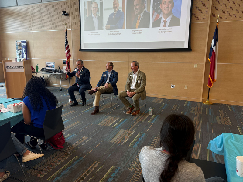 three men in chairs talking to group in big room