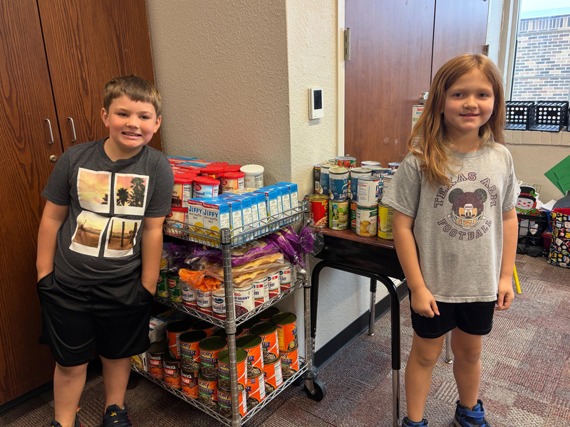 elememtary age boy and girl standing by cart full of canned goods